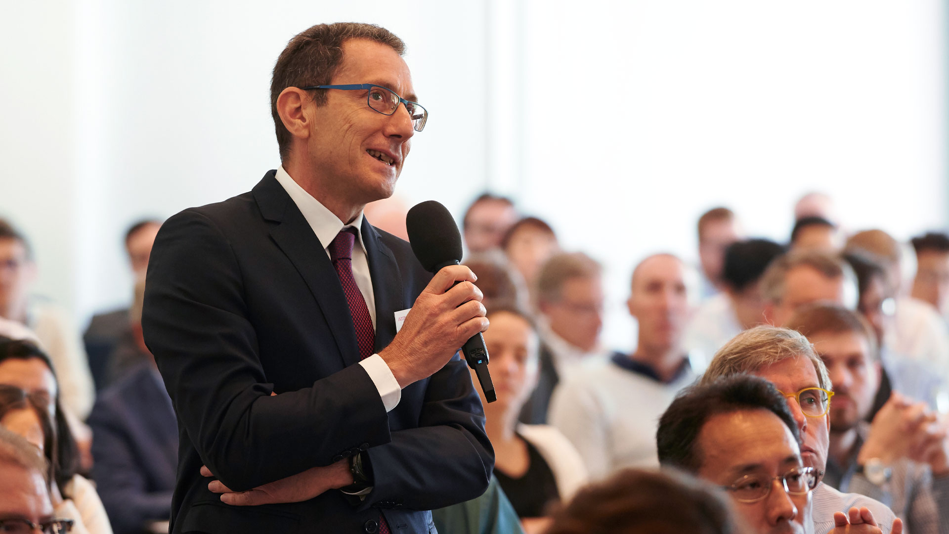 Frage aus dem Publikum. Mann mit Mikrofon stehend, umrandet von Plenum.  