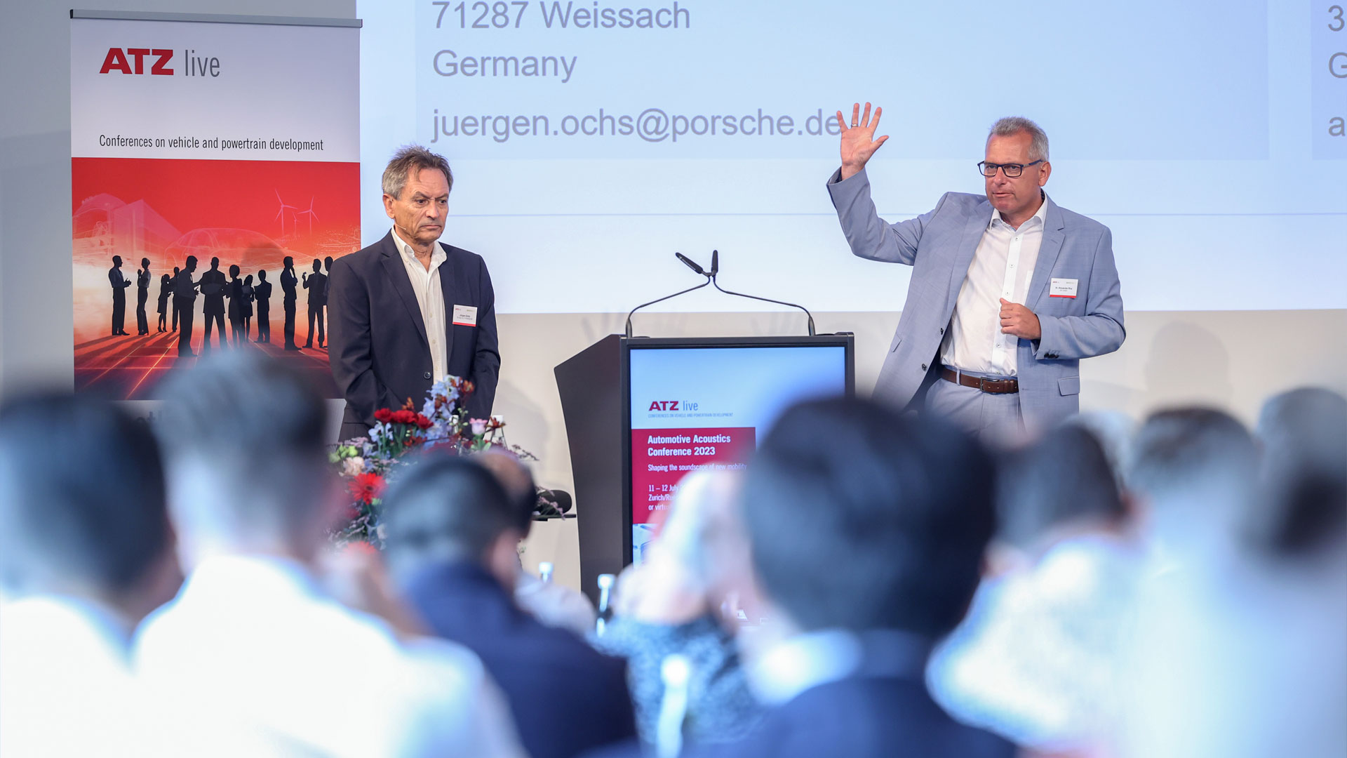 Zwei Referenten während ihres Vortrags auf der Bühne. Im Vordergrund das Publikum.