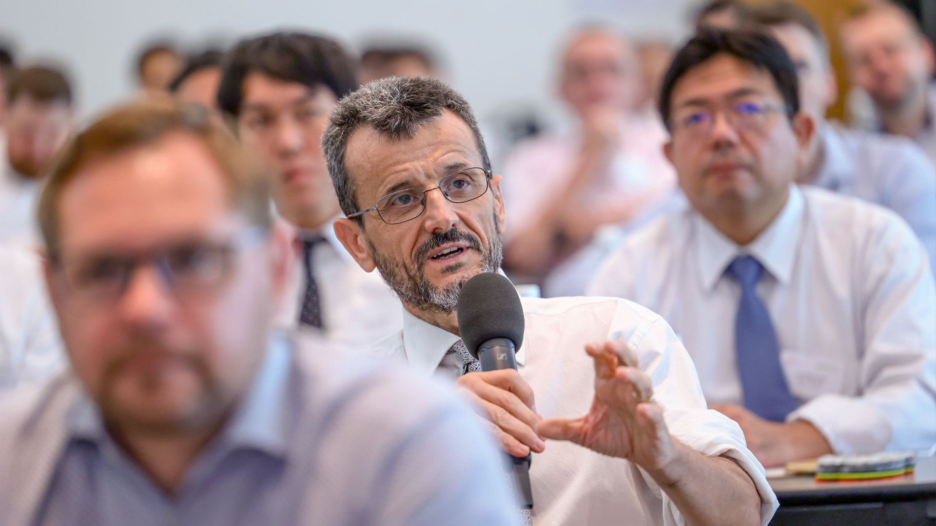 Frage aus dem Publikum. Mann mit Mikrofon sitzend, umrandet von Plenum.  
