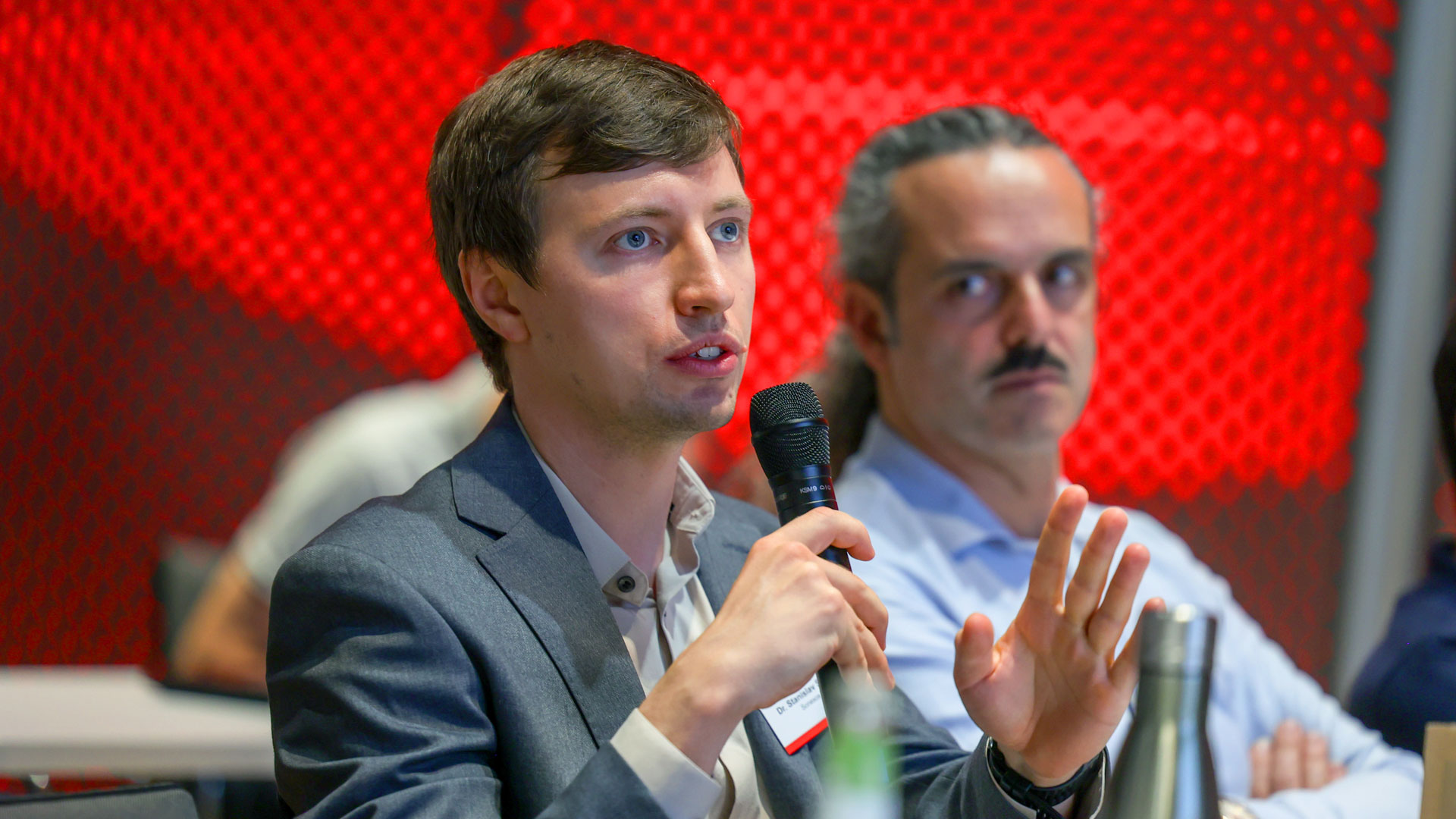 Frage aus dem Publikum. Mann mit Mikrofon sitzend, umrandet von Plenum.  