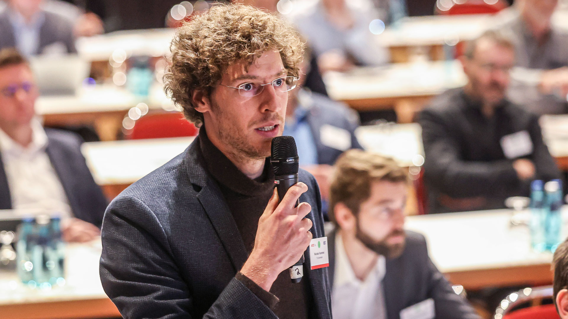 Question from the audience. Man with microphone standing, surrounded by plenum.