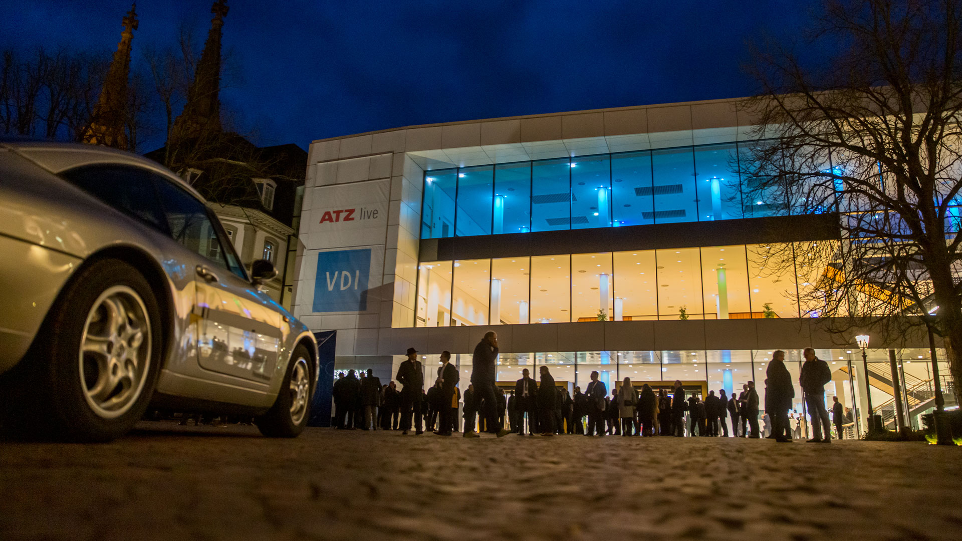Many conference participants in front of the congress center in the evening. In the foreground an exhibition vehicle. 